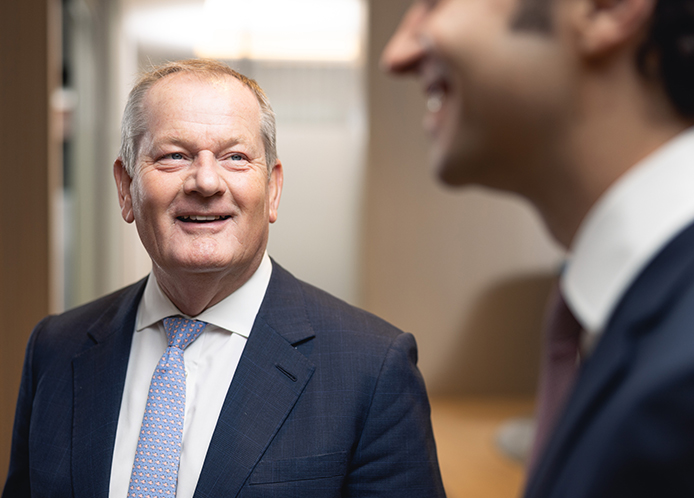 Man in suit smiles while looking at a younger man in suit in a professional setting.