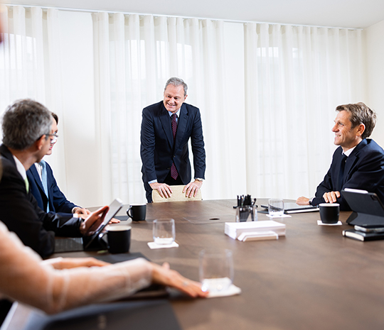 Four men in suits around a table in a meeting room, smiling and conversing.